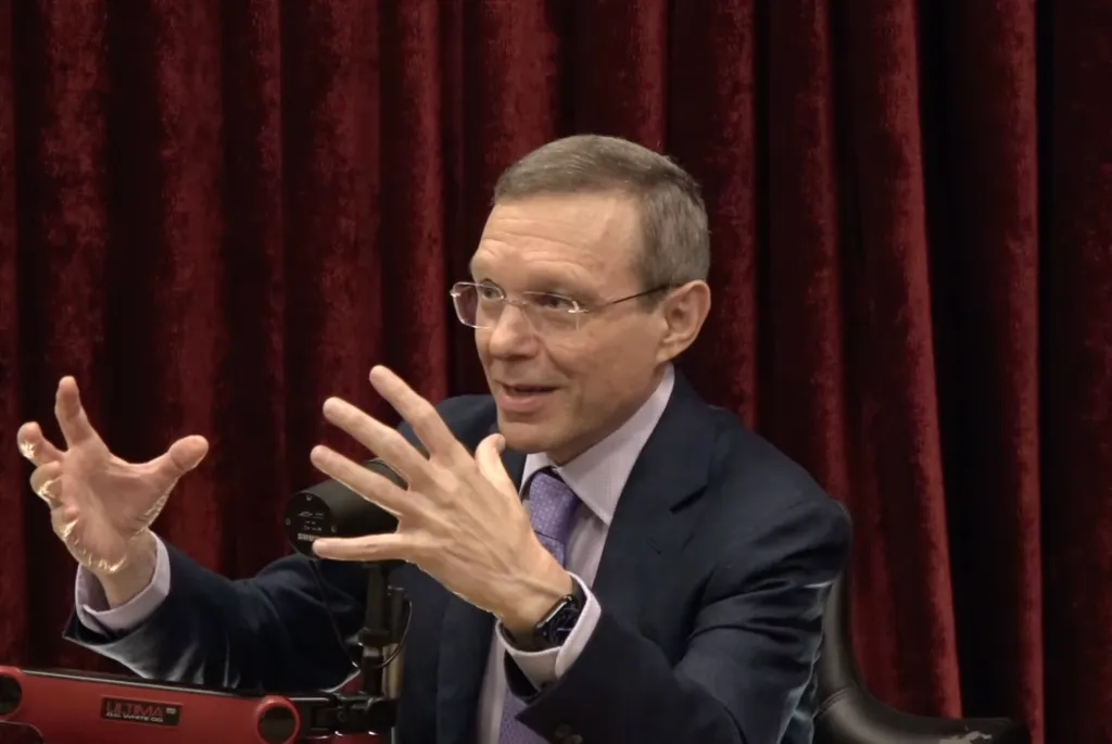 Man with glasses speaking, gesturing with both hands, in front of a red curtain.